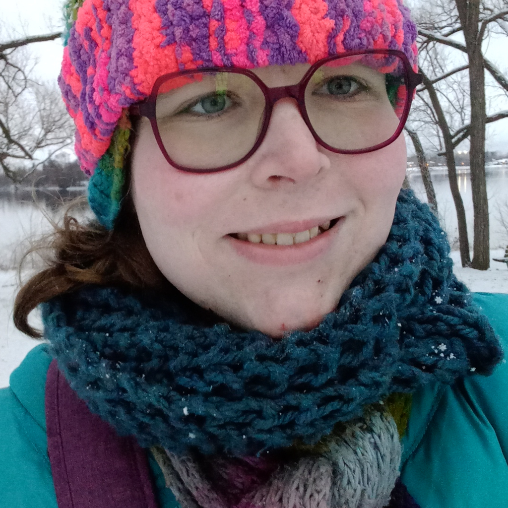 A joyful, smiling trans woman, with long, brown hair, standing in a snow-covered park on a cold day. There are also several trees, a park bench, and a lake in the background behind her. She is wearing several colorful hats, several colorful scarfs, and eyeglasses with purple frames.